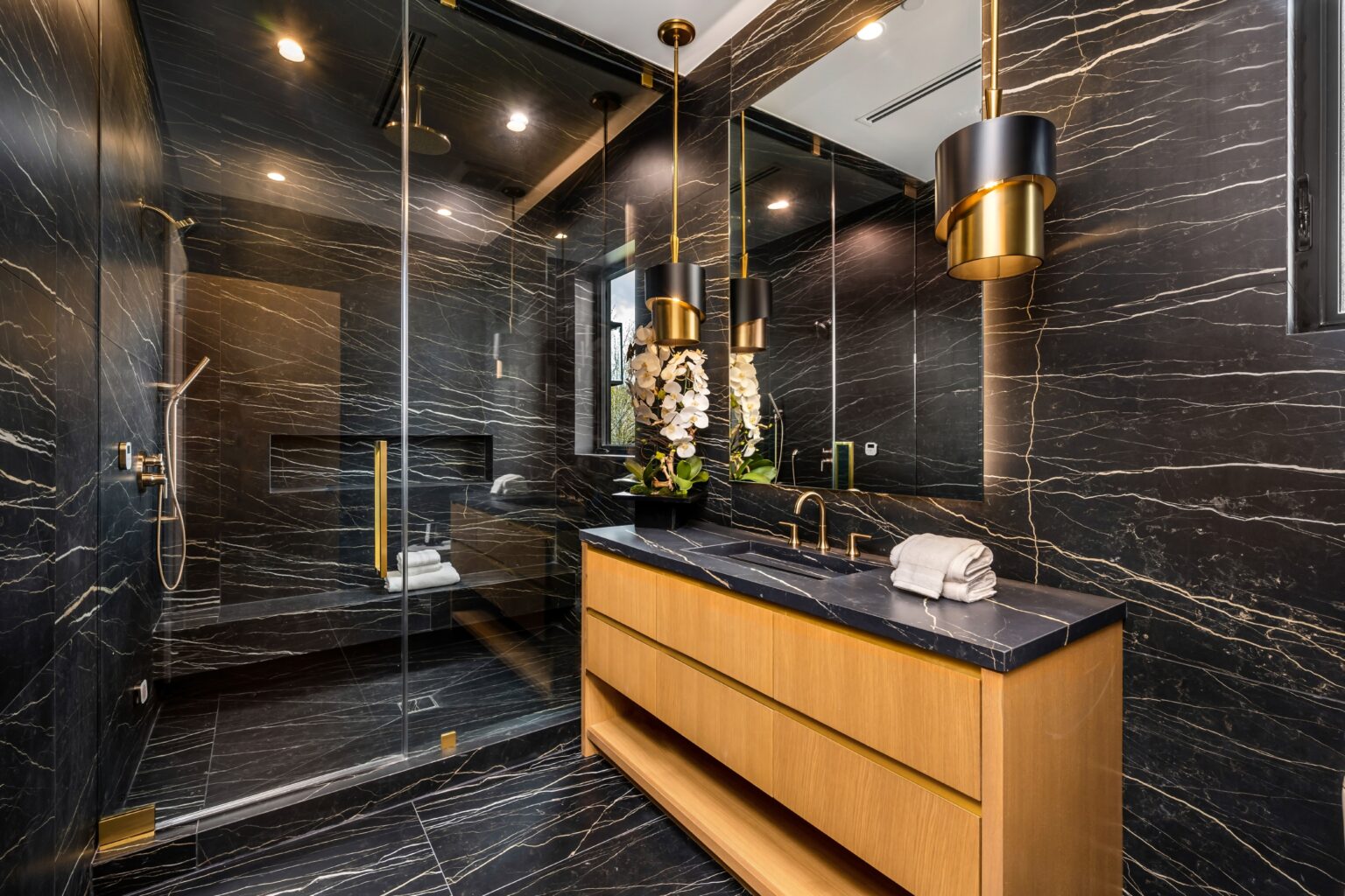 A luxurious modern bathroom with black marble walls, a glass shower, and a wooden vanity with gold accents.